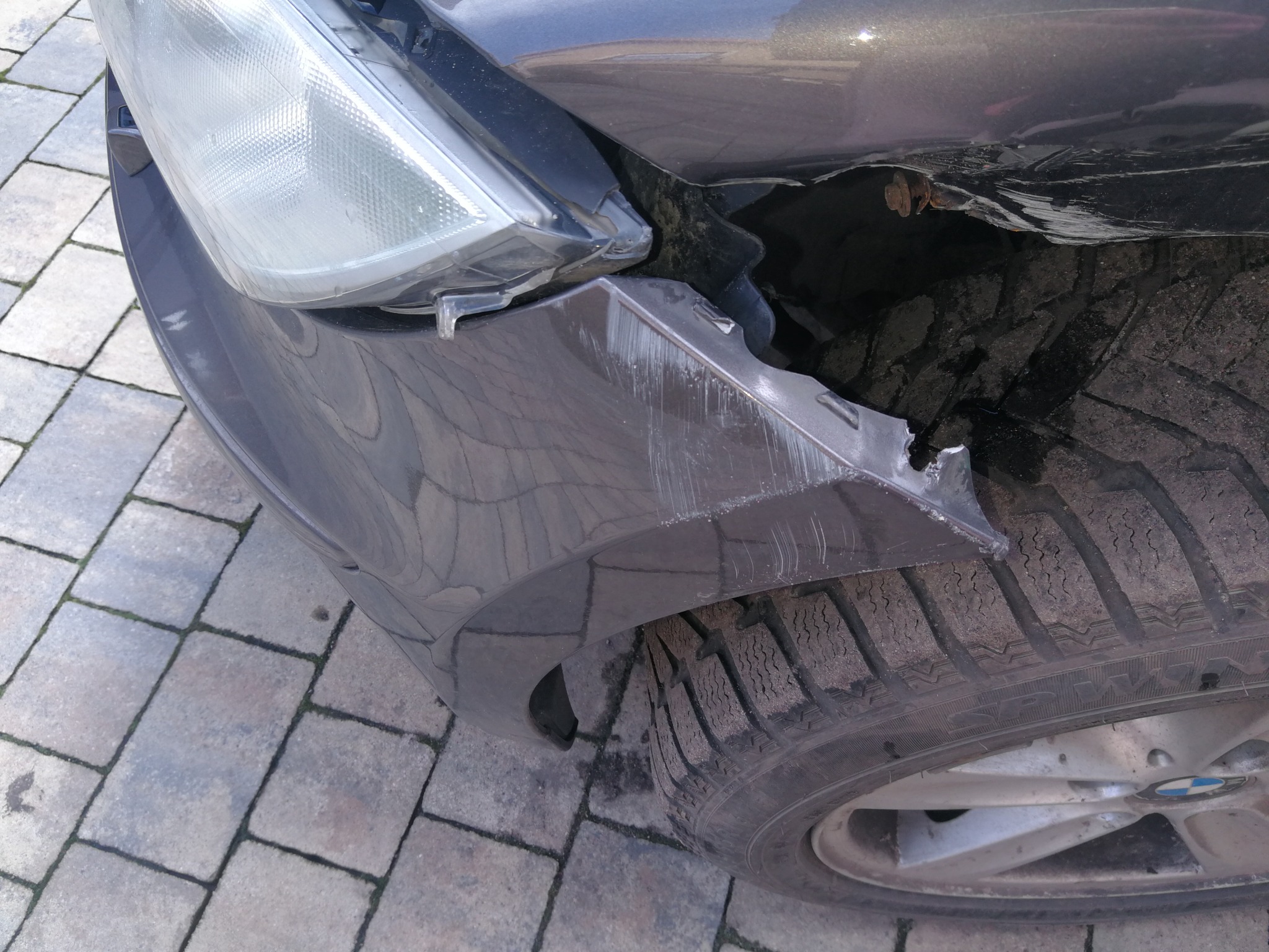 Uszkodzony przód samochodu: pęknięty zderzak, zarysowania i odsłonięte elementy konstrukcyjne przy kole. Widoczna opona zimowa i fragment felgi z logo BMW.