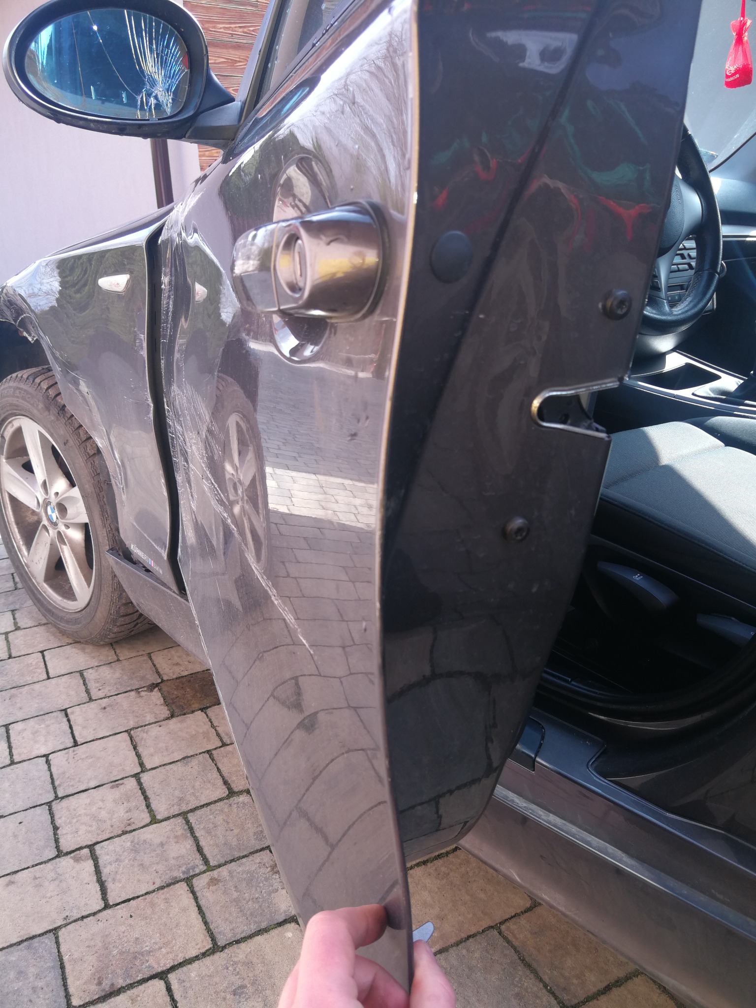 Uszkodzony, szary samochód marki BMW z pękniętym lusterkiem bocznym i wgnieceniami na drzwiach, stojący na brukowanej nawierzchni.