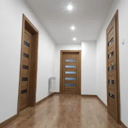 Wykończony korytarz z trzema drewnianymi drzwiami z szybami, podłoga z paneli imitujących drewno, białe ściany i sufit z oświetleniem punktowym.