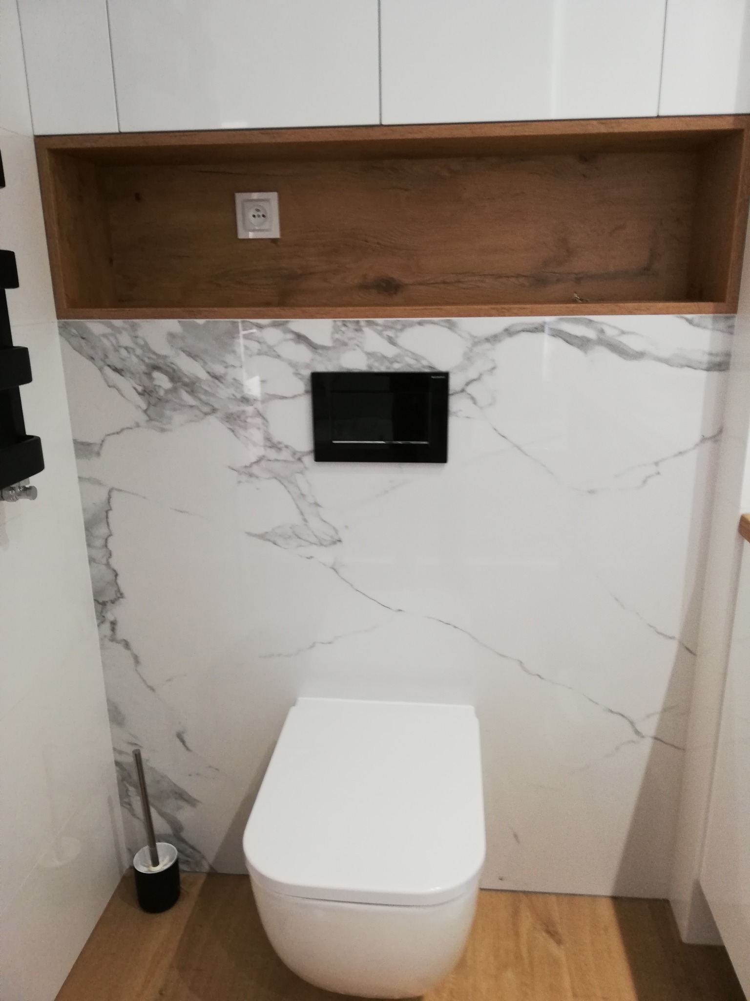 Nowoczesna toaleta podwieszana z białym sedesem i czarnym przyciskiem spłukującym na tle marmurowych płytek, nad nią drewniana wnęka z gniazdkiem elektrycznym, widoczna szczotka do WC.
