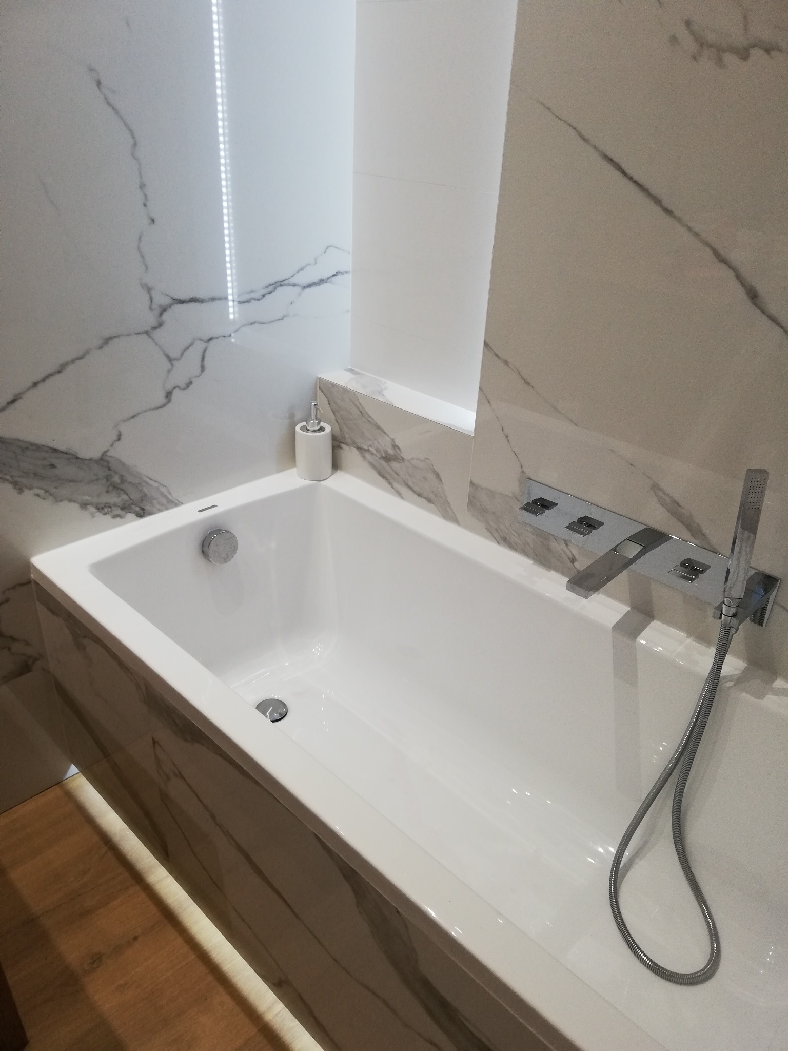 Nowoczesna, prostokątna wanna obudowana płytkami imitującymi marmur, z chromowaną armaturą i słuchawką prysznicową, w łazience wykończonej płytkami z motywem marmuru.