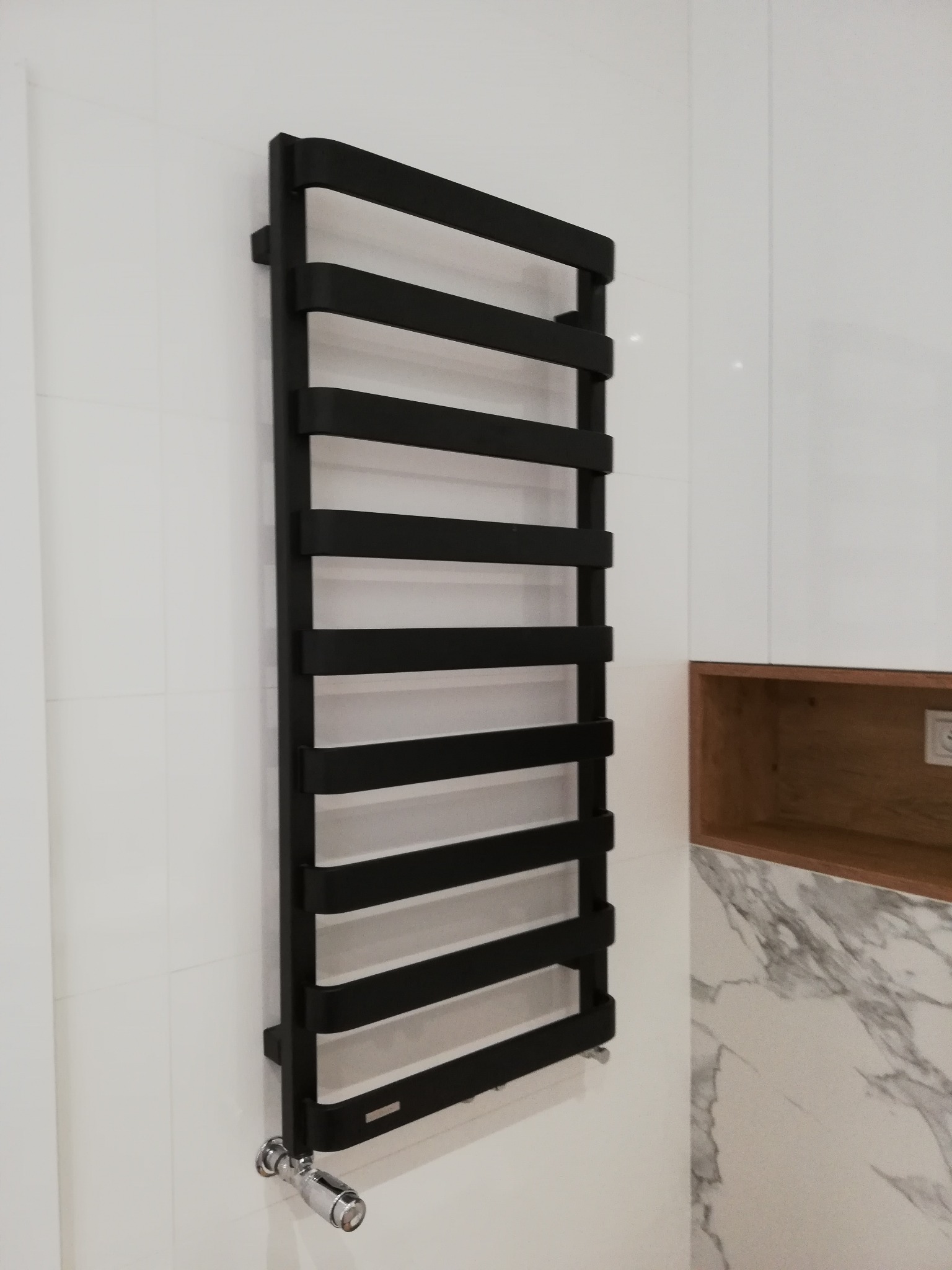 Czarny, drabinkowy grzejnik łazienkowy na białej ścianie z jasnymi płytkami, widoczny fragment marmurowego wykończenia i drewniana półka.