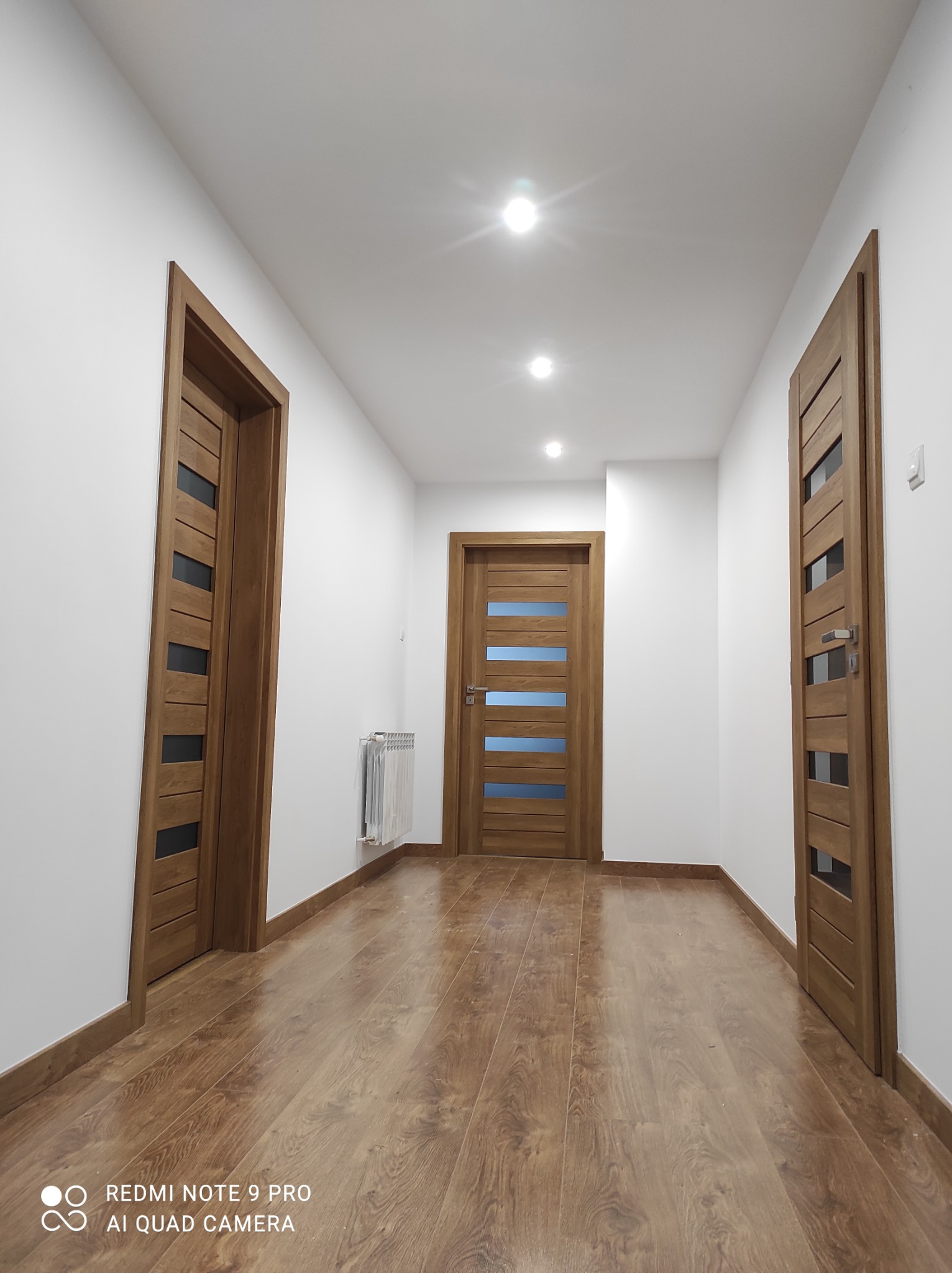 Wykończony korytarz z trzema drewnianymi drzwiami z szybami, podłoga z paneli imitujących drewno, białe ściany i sufit z oświetleniem punktowym.