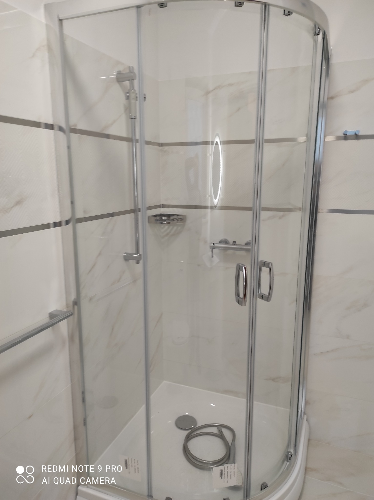 Kabina prysznicowa z chromowanymi profilami i drzwiami przesuwnymi, zamontowana w łazience wyłożonej białymi płytkami imitującymi marmur z szarymi paskami dekoracyjnymi, widoczny wąż prysznicowy...