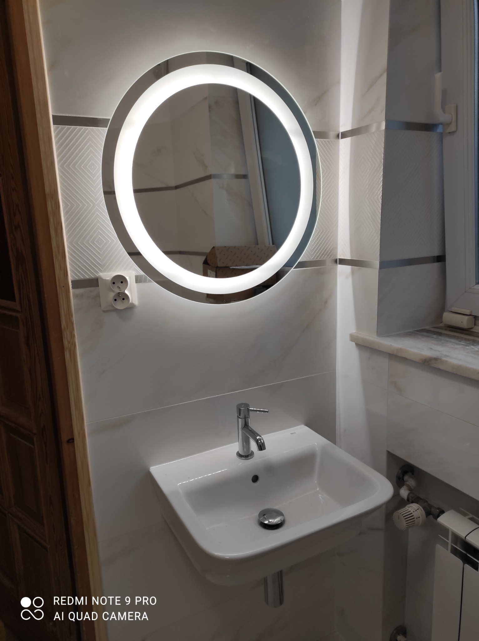 Okrągłe lustro z podświetleniem LED nad białą umywalką, marmurowe płytki na ścianie z widocznym gniazdkiem elektrycznym i grzejnikiem pod oknem.