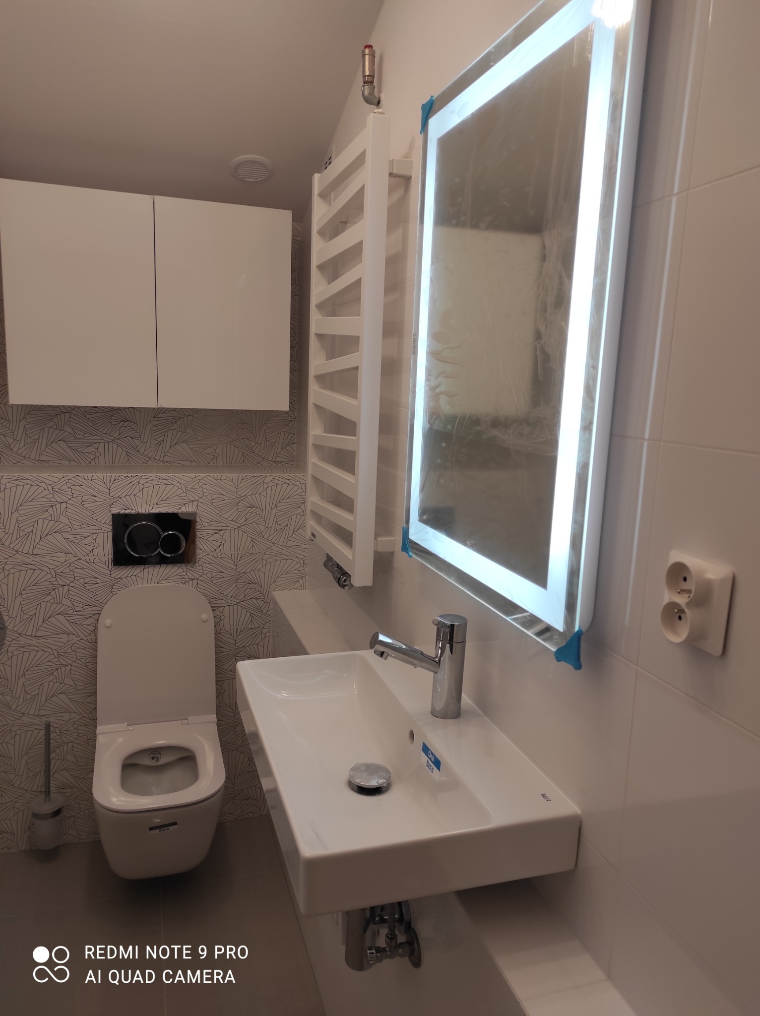 Nowoczesna łazienka z podświetlanym lustrem, wiszącą toaletą i umywalką Roca, grzejnik drabinkowy oraz wzorzyste płytki na ścianie za toaletą.