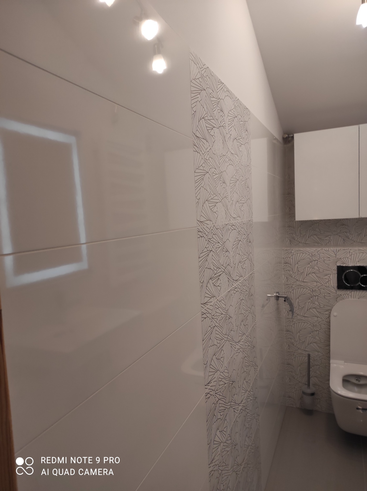 Białe, błyszczące płytki ścienne ułożone poziomo sąsiadują z dekoracyjnymi płytkami ze wzorem w odcieniach szarości w wyremontowanej łazience. Widoczna toaleta i szafka wisząca.