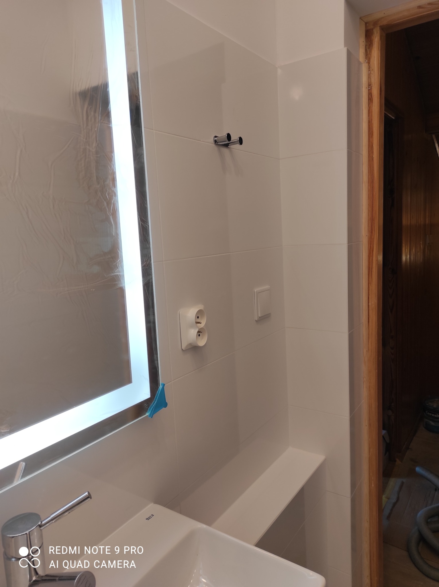 Biała łazienka z umywalką, lustrem z podświetleniem LED w folii ochronnej, gładkimi białymi płytkami, drewnianą ościeżnicą i widocznym gniazdkiem elektrycznym oraz włącznikiem światła.