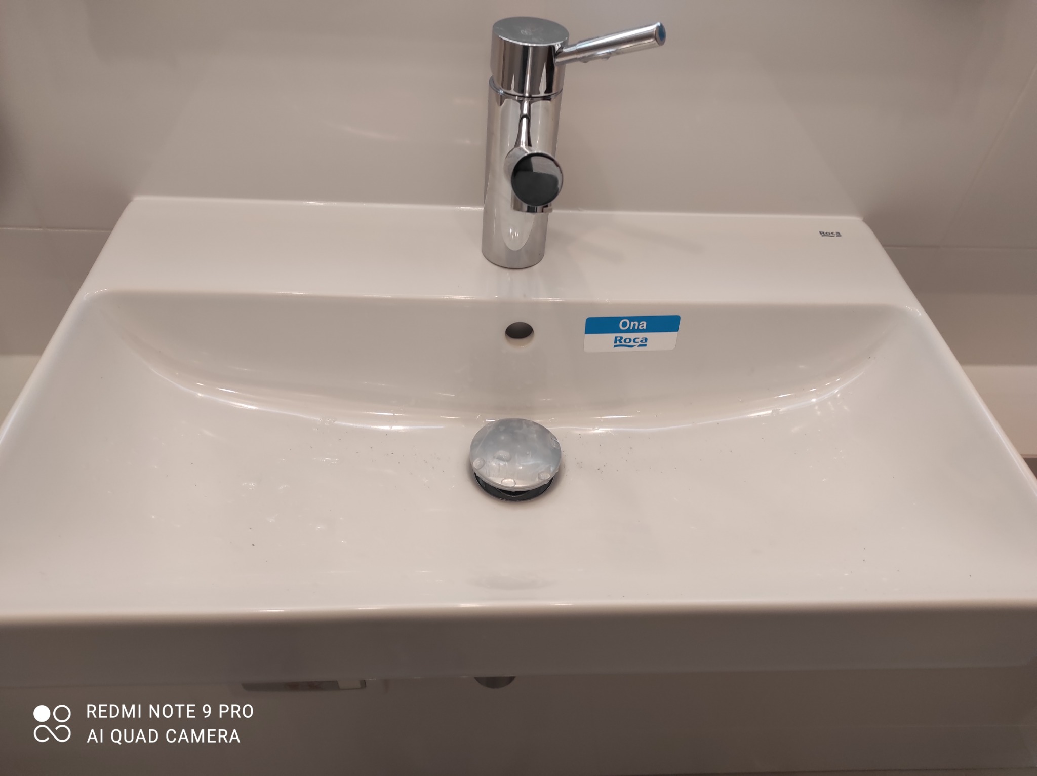 Biała umywalka Roca Ona z chromowaną baterią i korkiem, zamontowana na tle jasnej ściany.