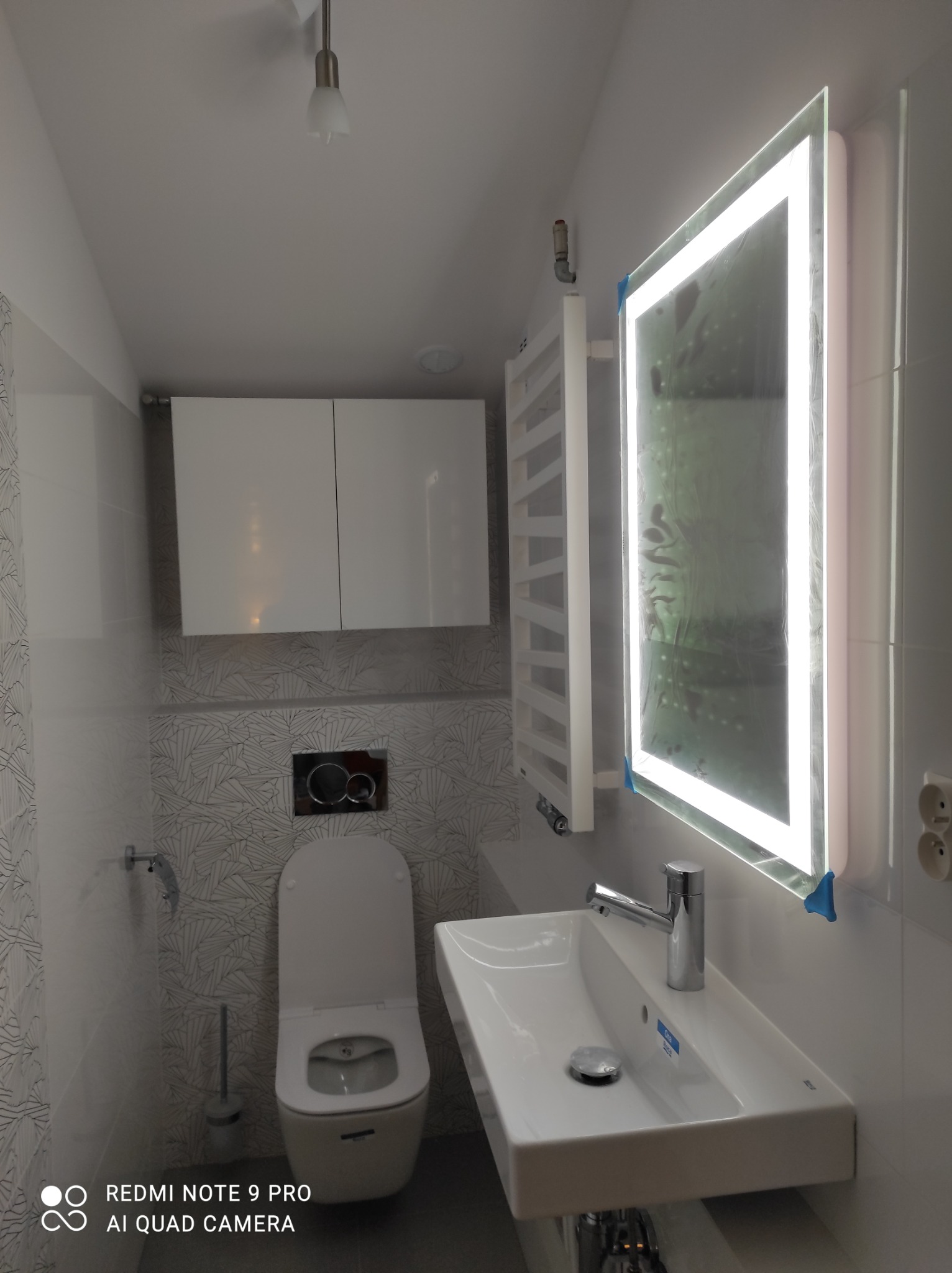 Nowoczesna łazienka po remoncie: podwieszana toaleta, umywalka z podświetlanym lustrem, grzejnik drabinkowy i szafki wiszące na tle wzorzystych płytek.