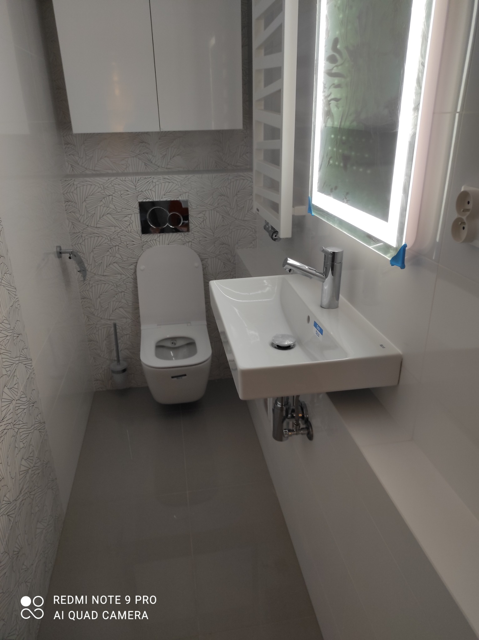 Nowoczesna łazienka z podwieszaną toaletą, prostokątną umywalką z chromowaną baterią i podświetlanym lustrem, wykończona płytkami w odcieniach szarości i bieli.