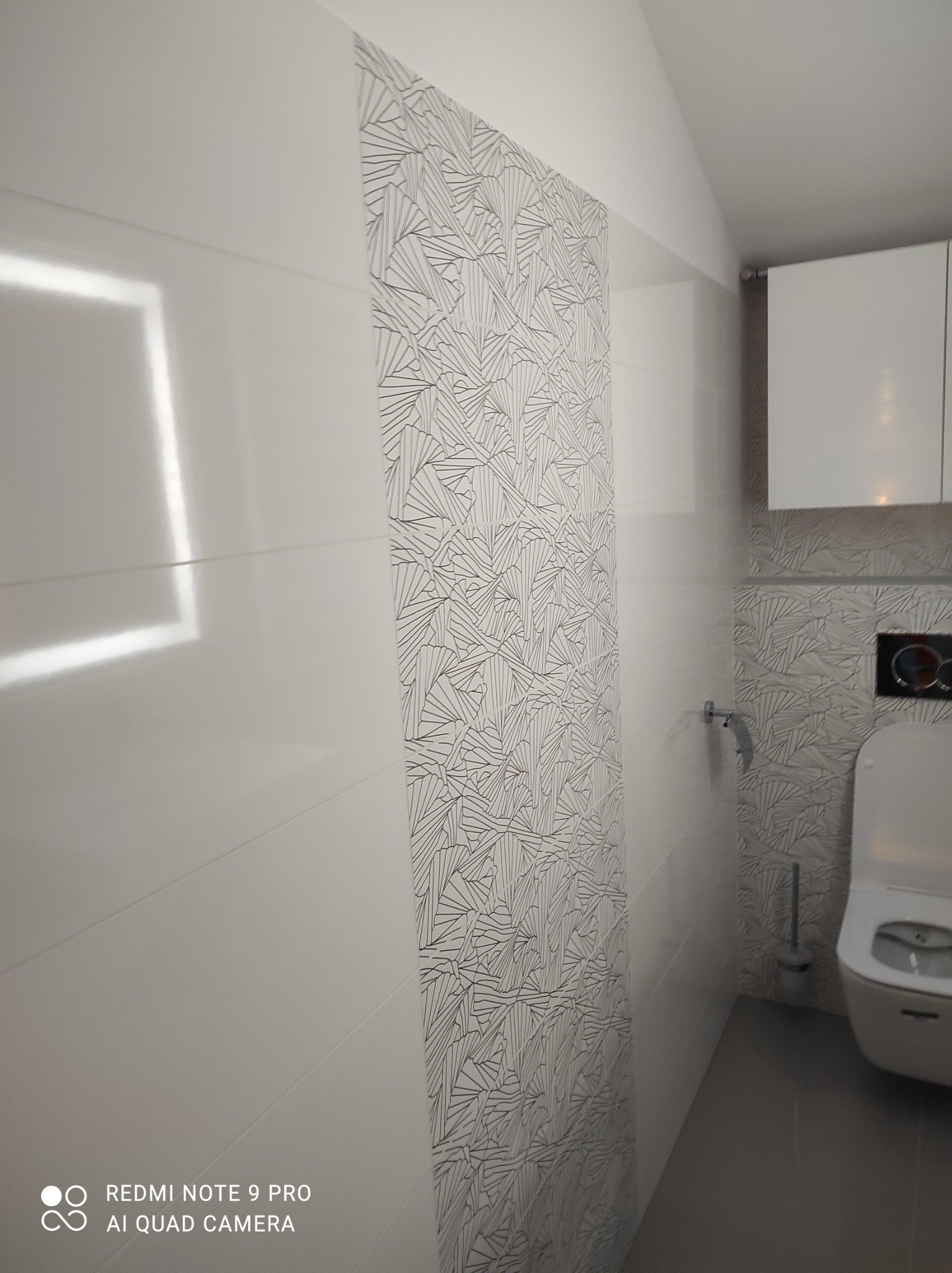 Biała łazienka z płytkami w geometryczny wzór na ścianie, wisząca toaleta i szafka nad nią, widoczne odbicie światła na gładkich płytkach.