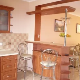 Drewniany barek kuchenny z chromowanym stojakiem na kieliszki i owocami, z dwoma chromowanymi krzesłami barowymi na tle jasnej kuchni i salonu.