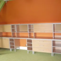 Zabudowa meblowa na wymiar z jasnego drewna, z szufladami, szafkami i otwartymi półkami, na tle pomarańczowej ściany i zielonego filaru konstrukcyjnego.