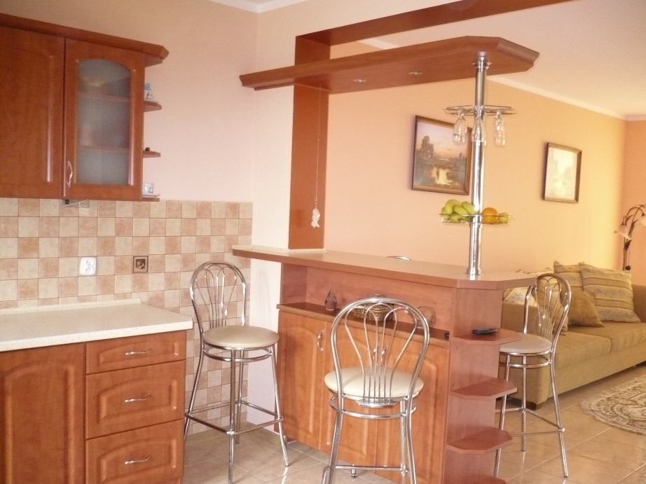 Drewniany barek kuchenny z chromowanym stojakiem na kieliszki i owocami, z dwoma chromowanymi krzesłami barowymi na tle jasnej kuchni i salonu.