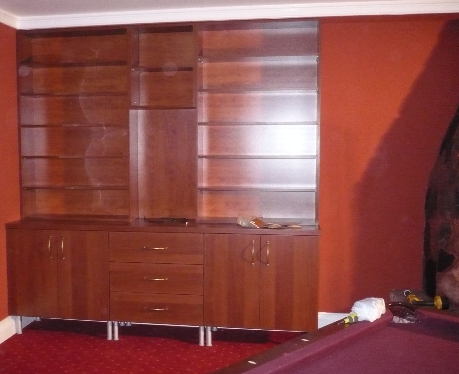 Wieloczęściowa, drewniana szafa wnękowa z półkami i szafkami, z widocznymi srebrnymi nóżkami, umieszczona na tle czerwonej ściany i czerwonej wykładziny podłogowej, z elementami montażowymi na blacie.