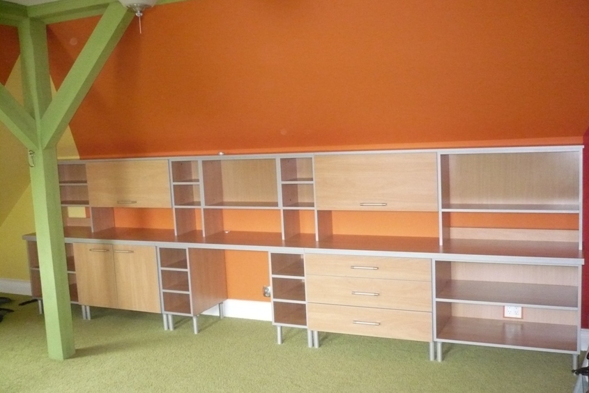 Zabudowa meblowa na wymiar z jasnego drewna, z szufladami, szafkami i otwartymi półkami, na tle pomarańczowej ściany i zielonego filaru konstrukcyjnego.