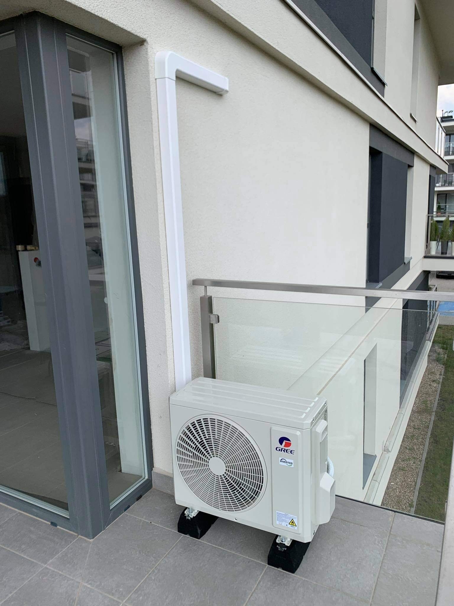 Zewnętrzna jednostka klimatyzacyjna marki Gree zamontowana na balkonie nowoczesnego apartamentu, widoczne białe rury odprowadzające skropliny i czarne podkładki antywibracyjne pod urządzeniem.