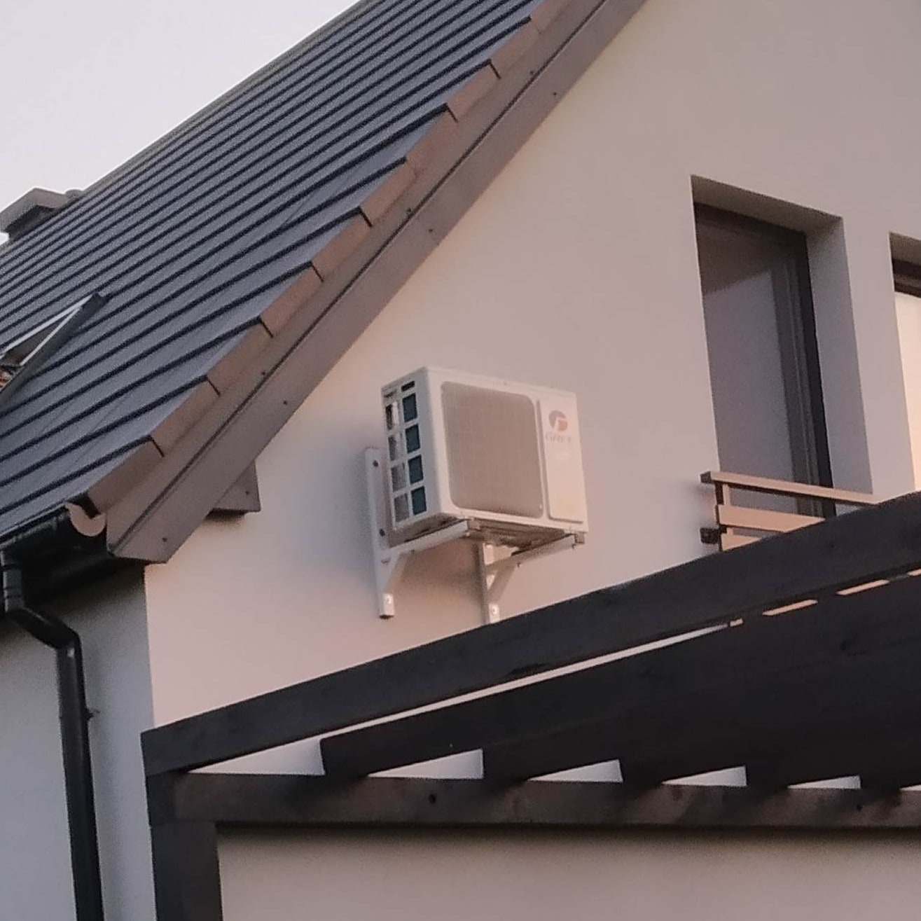 Biała jednostka zewnętrzna klimatyzatora zamontowana na ścianie domu z ciemną dachówką, widoczna z perspektywy spod pergoli.