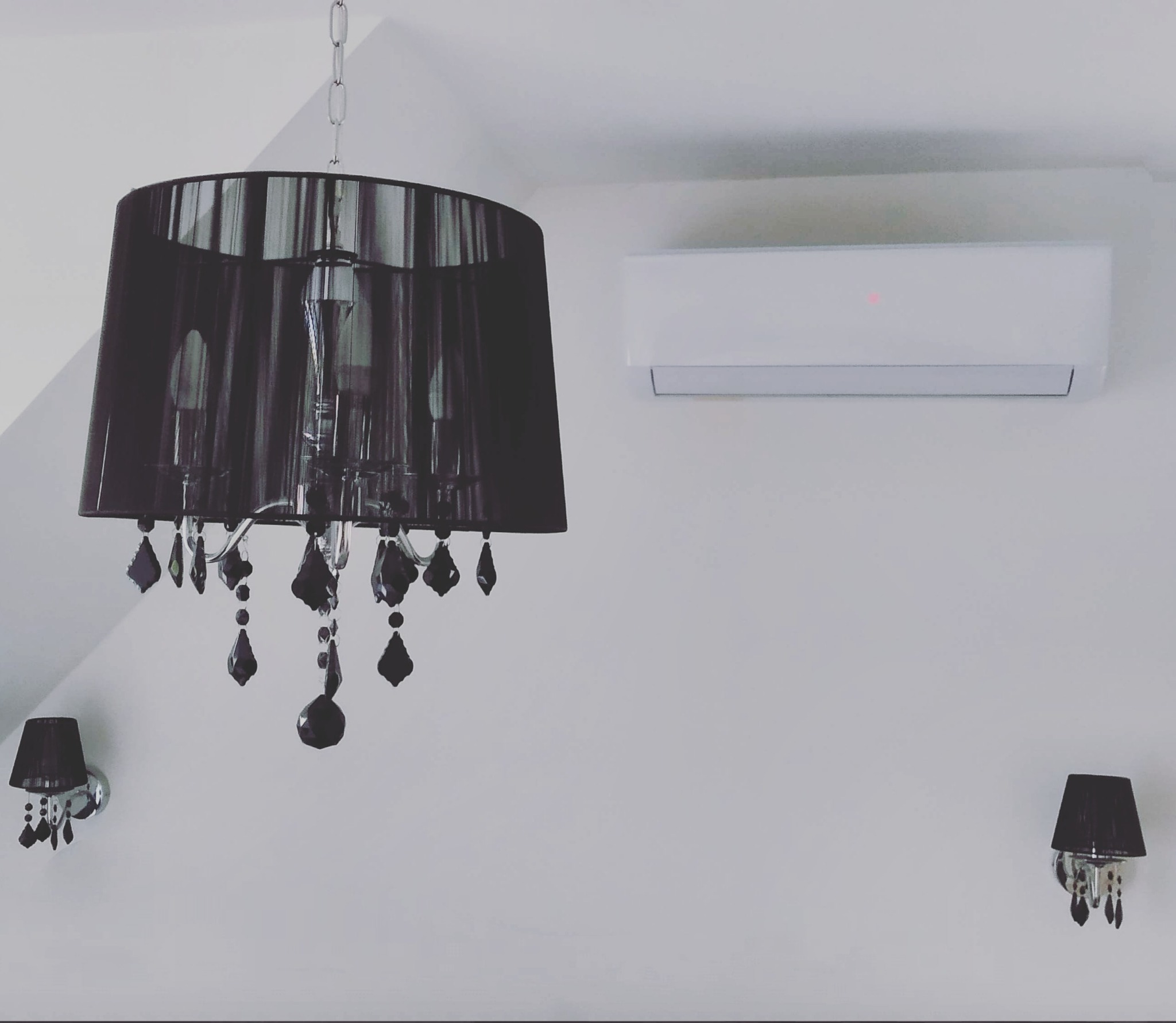 Biały klimatyzator ścienny z czerwoną diodą, zawieszony nad czarnym, kryształowym żyrandolem i dwoma kinkietami z czarnymi abażurami na białej ścianie.