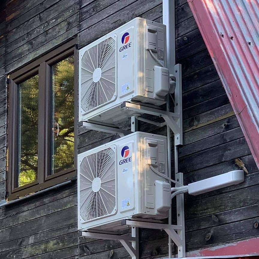 Dwa klimatyzatory marki Gree zamontowane na zewnątrz drewnianego domu z widocznym oknem i fragmentem dachu.