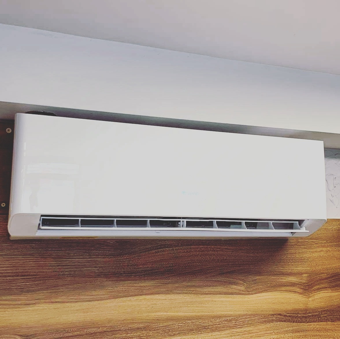 Biała jednostka wewnętrzna klimatyzatora zamontowana na ścianie z panelami drewnopodobnymi, widoczne otwory wentylacyjne i subtelne logo producenta.