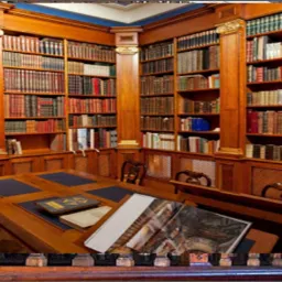 Drewniana biblioteka z rzędami książek, biurkiem z otwartą księgą, prezentująca wnętrze gabinetu.