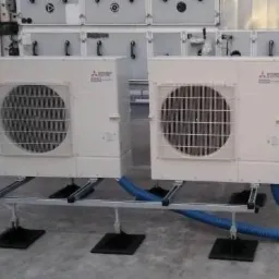 Dwie zewnętrzne jednostki klimatyzacyjne Mitsubishi Electric na dachu, zamontowane na metalowych konstrukcjach wsporczych z niebieskimi przewodami. Widoczne elementy wentylacyjne VTS w tle.