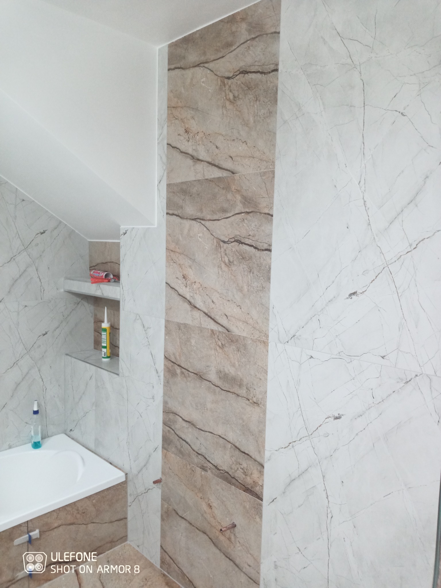 Wykończona łazienka z marmurowymi płytkami w odcieniach bieli i beżu, widoczna półka z silikonem i kosmetykami, oraz fragment umywalki z dozownikiem mydła.