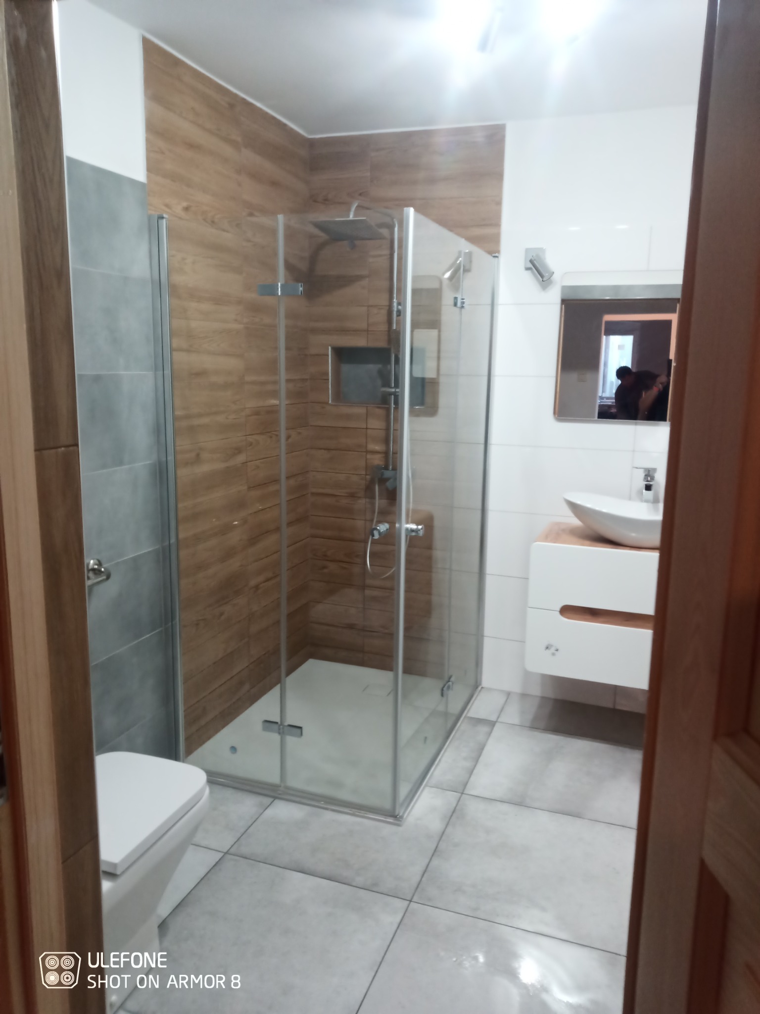 Nowoczesna łazienka po remoncie z kabiną prysznicową typu walk-in z drewnopodobnymi płytkami, białą umywalką nablatową na szafce z drewnianym akcentem oraz toaletą.