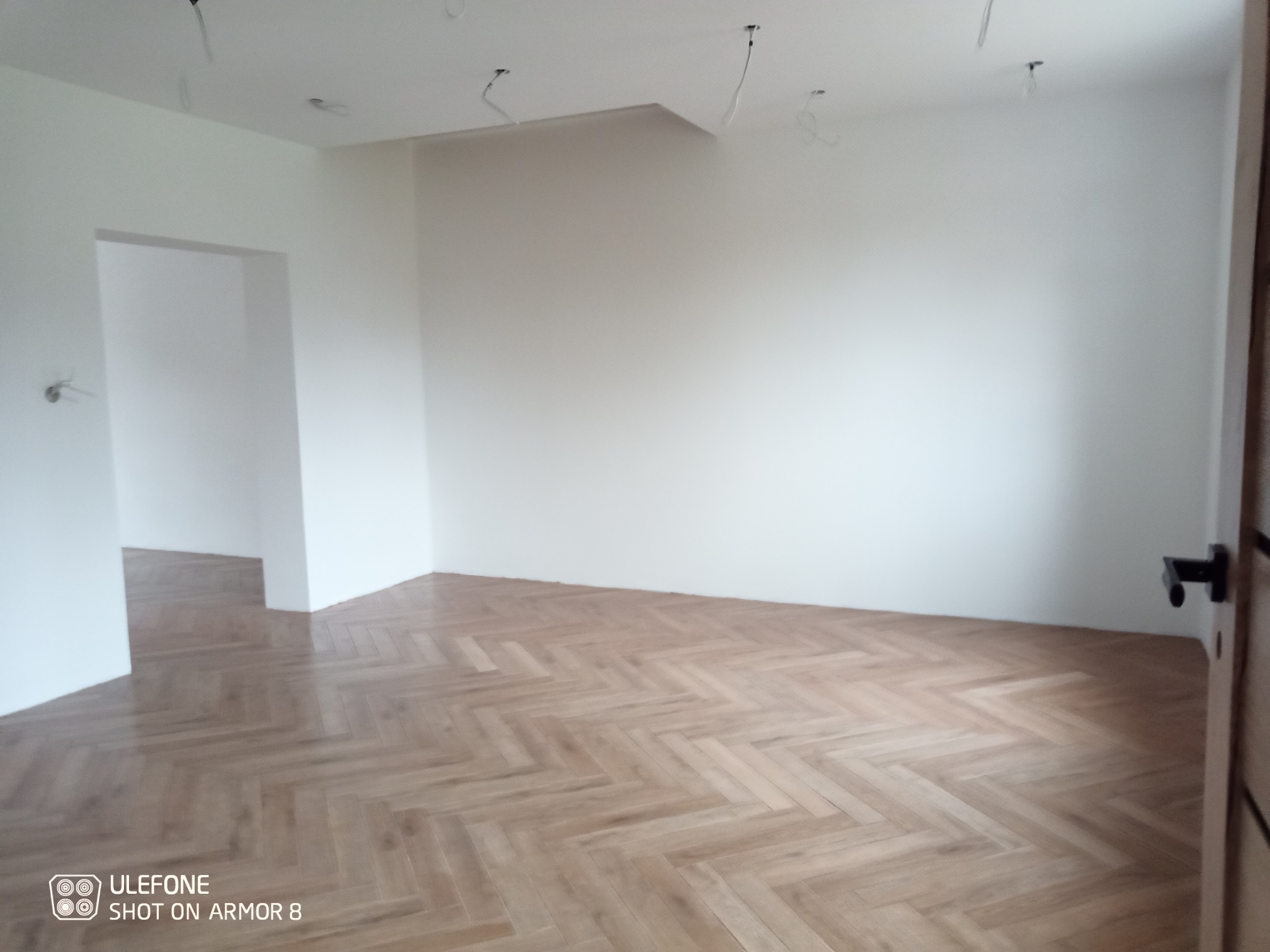 Pusty, odświeżony pokój z parkietem ułożonym w jodełkę, świeżo pomalowanymi białymi ścianami i widocznymi przewodami elektrycznymi wystającymi z sufitu.