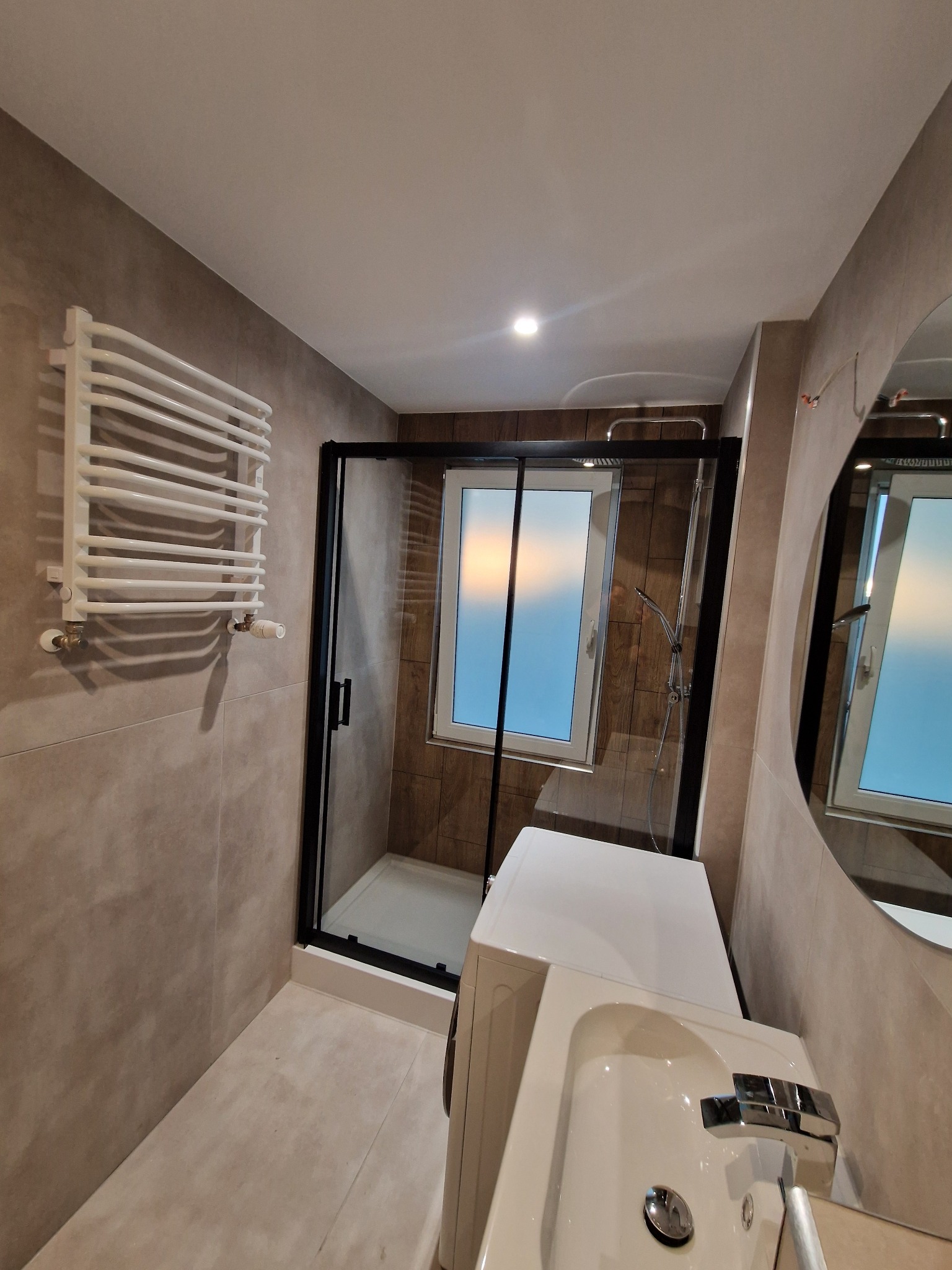 Wykończona łazienka w beżach: kabina prysznicowa z czarną ramą, grzejnik drabinkowy, umywalka z szafką i okrągłe lustro. Ukończony remont łazienki.