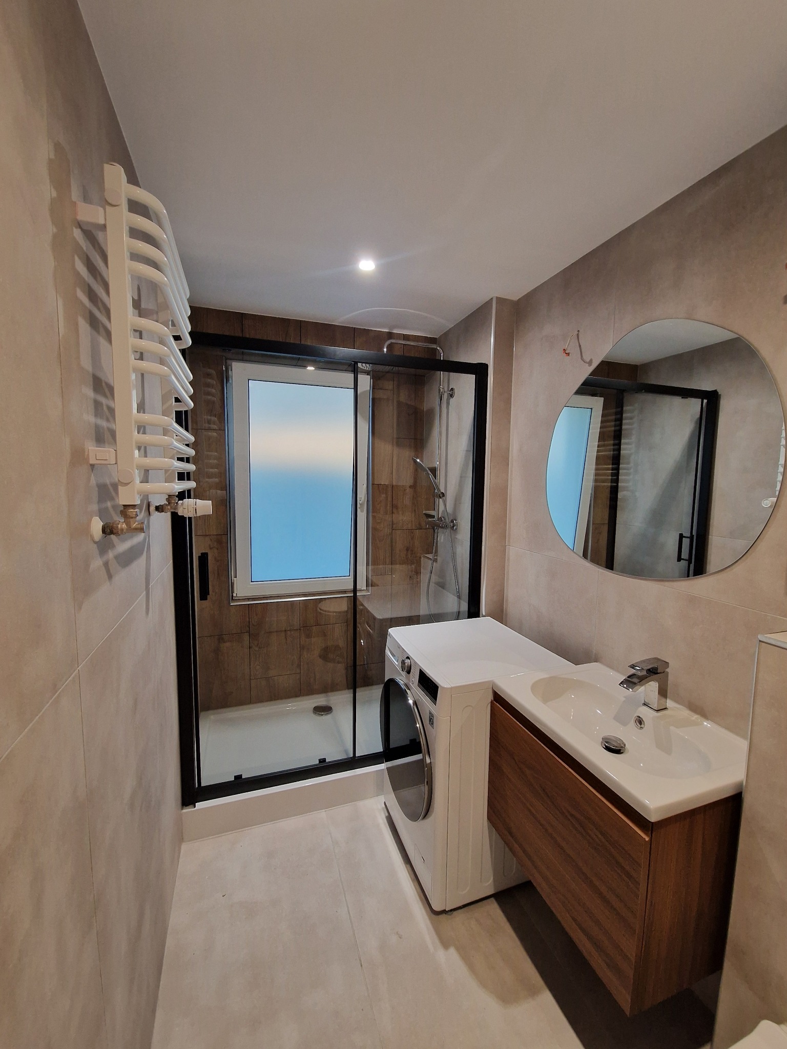 Nowoczesna łazienka po remoncie: kabina prysznicowa z czarną ramą, pralka obok umywalki z szafką z drewna, okrągłe lustro i grzejnik drabinkowy na beżowych płytkach.