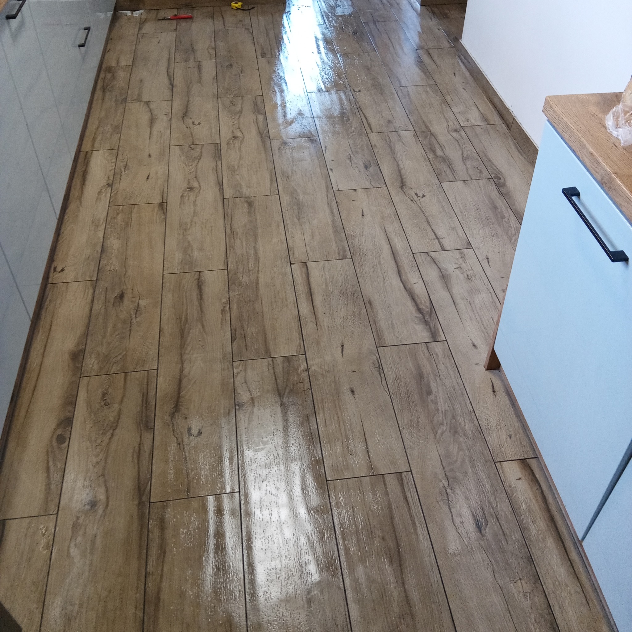 Ułożona podłoga z płytek imitujących drewno w kuchni, z widocznymi fragmentami szafek w kolorze błękitnym i narzędziami w tle. Błyszcząca powierzchnia płytek odbija światło.