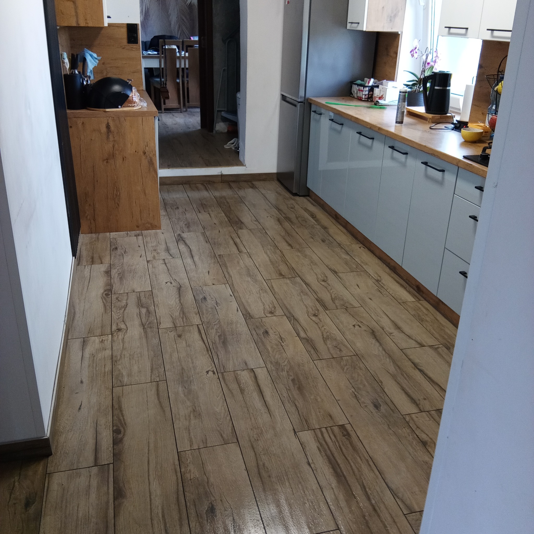 Ukończona podłoga w kuchni z drewnopodobnych płytek w Radzyniu Chełmińskim. Widoczne szafki kuchenne w tle i przejście do innego pomieszczenia.