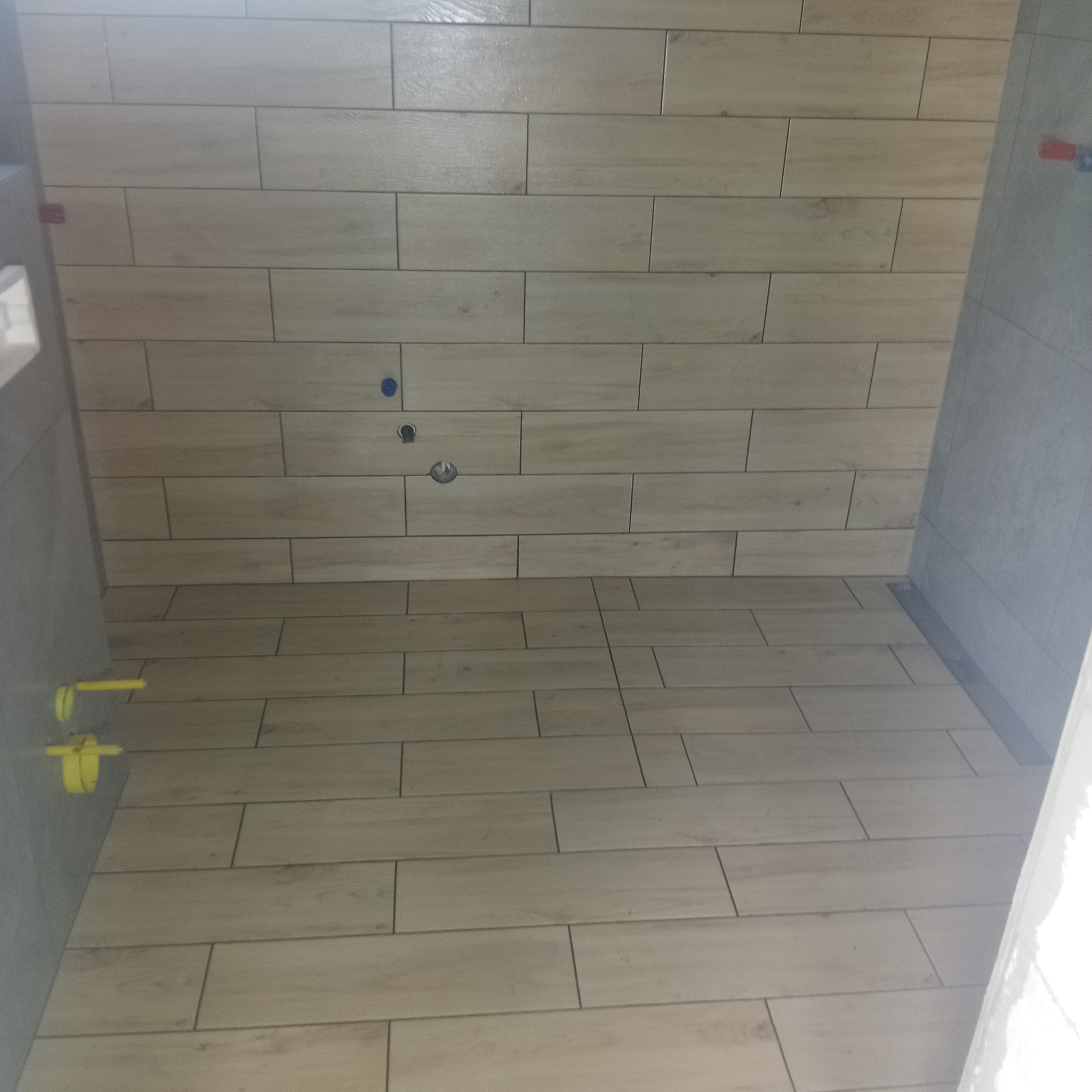 Łazienka po remoncie: jasne, podłużne płytki imitujące drewno na ścianach i podłodze, widoczne przyłącza wodne i odpływ liniowy. Nowoczesne wykończenie.