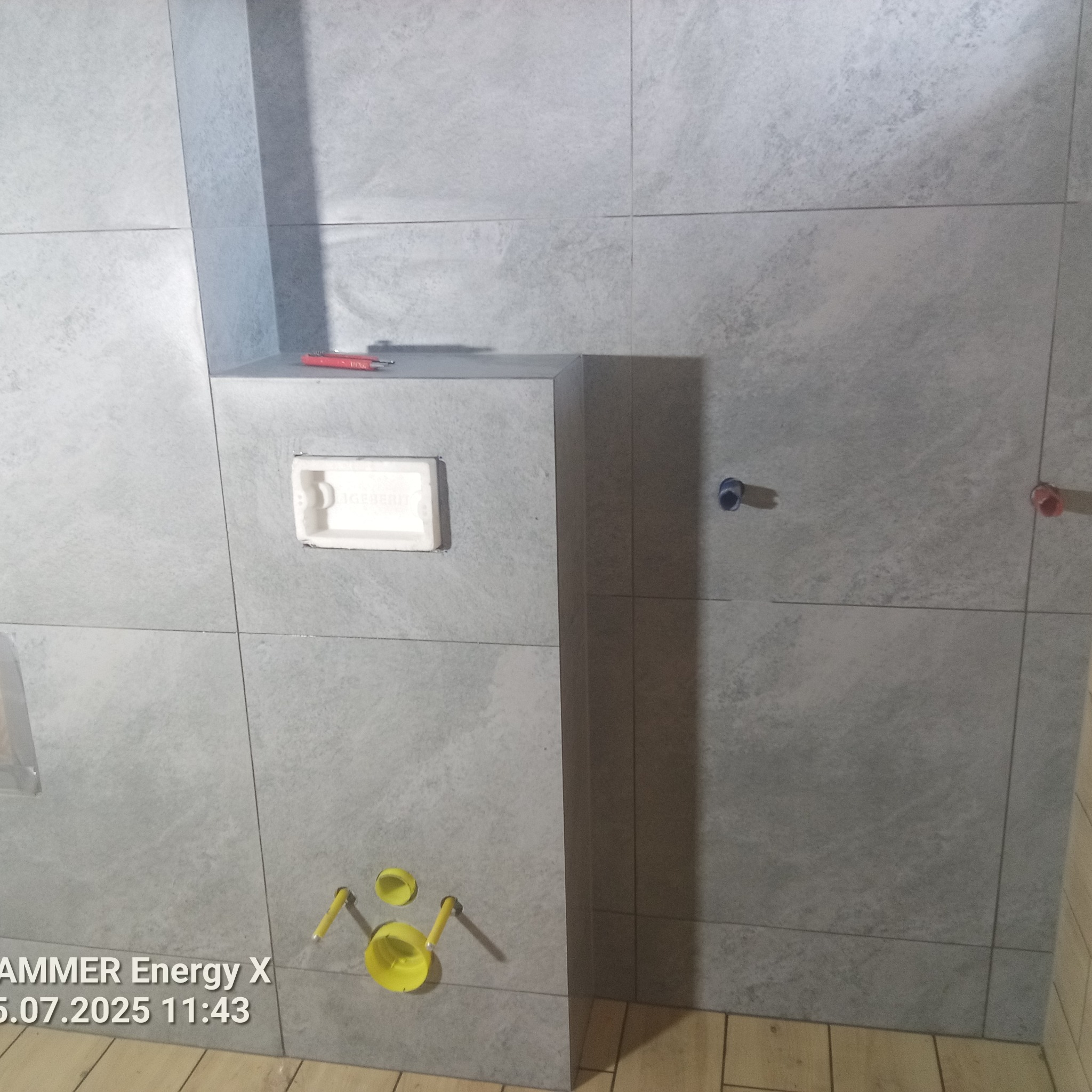 Szare płytki w łazience, zabudowa stelaża WC z widocznymi przyłączami wody i odpływu. Na zabudowie leży czerwony ołówek. Widoczny otwór rewizyjny.