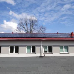 Panele słoneczne na dachu budynku z czerwonymi dachówkami, widok z poziomu dachu, błękitne niebo z chmurami w tle.