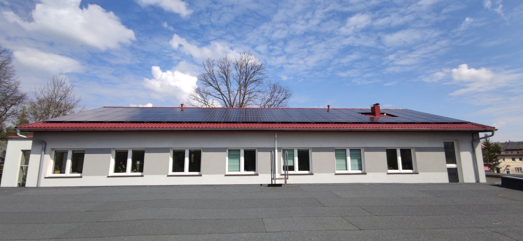 Panele słoneczne na dachu budynku z czerwonymi dachówkami, widok z poziomu dachu, błękitne niebo z chmurami w tle.