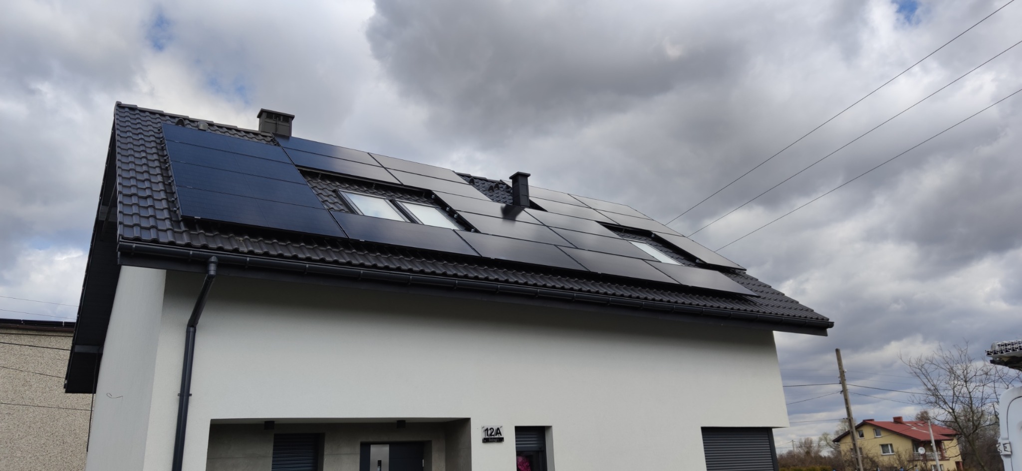 Czarny system paneli słonecznych na ciemnym dachu domu jednorodzinnego z białym wykończeniem, widoczne okna dachowe i pochmurne niebo.