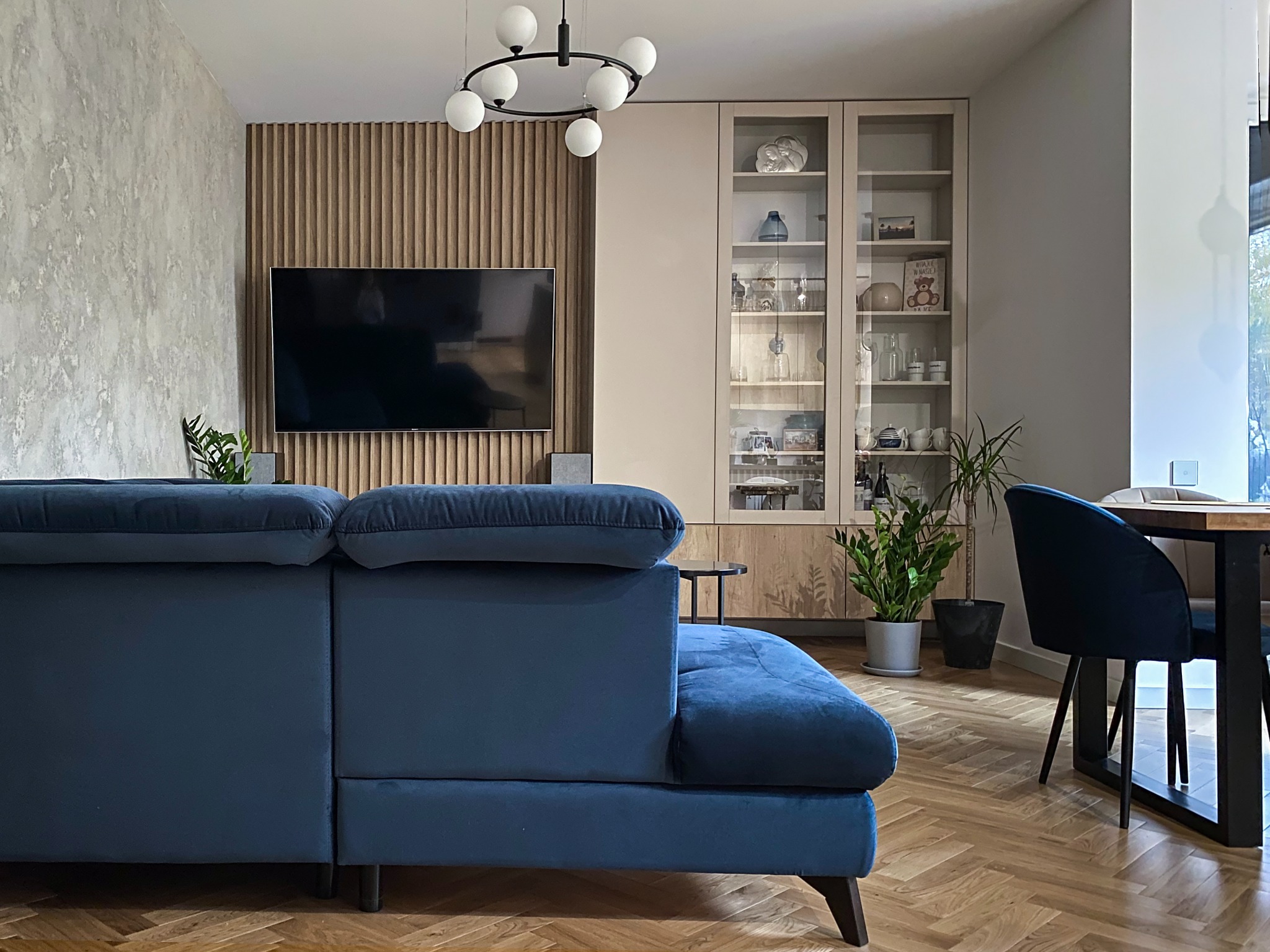 Salon z niebieską sofą, drewnianą ścianą z telewizorem, przeszkloną witryną, parkietem w jodełkę i nowoczesną lampą sufitową z białymi kulami.