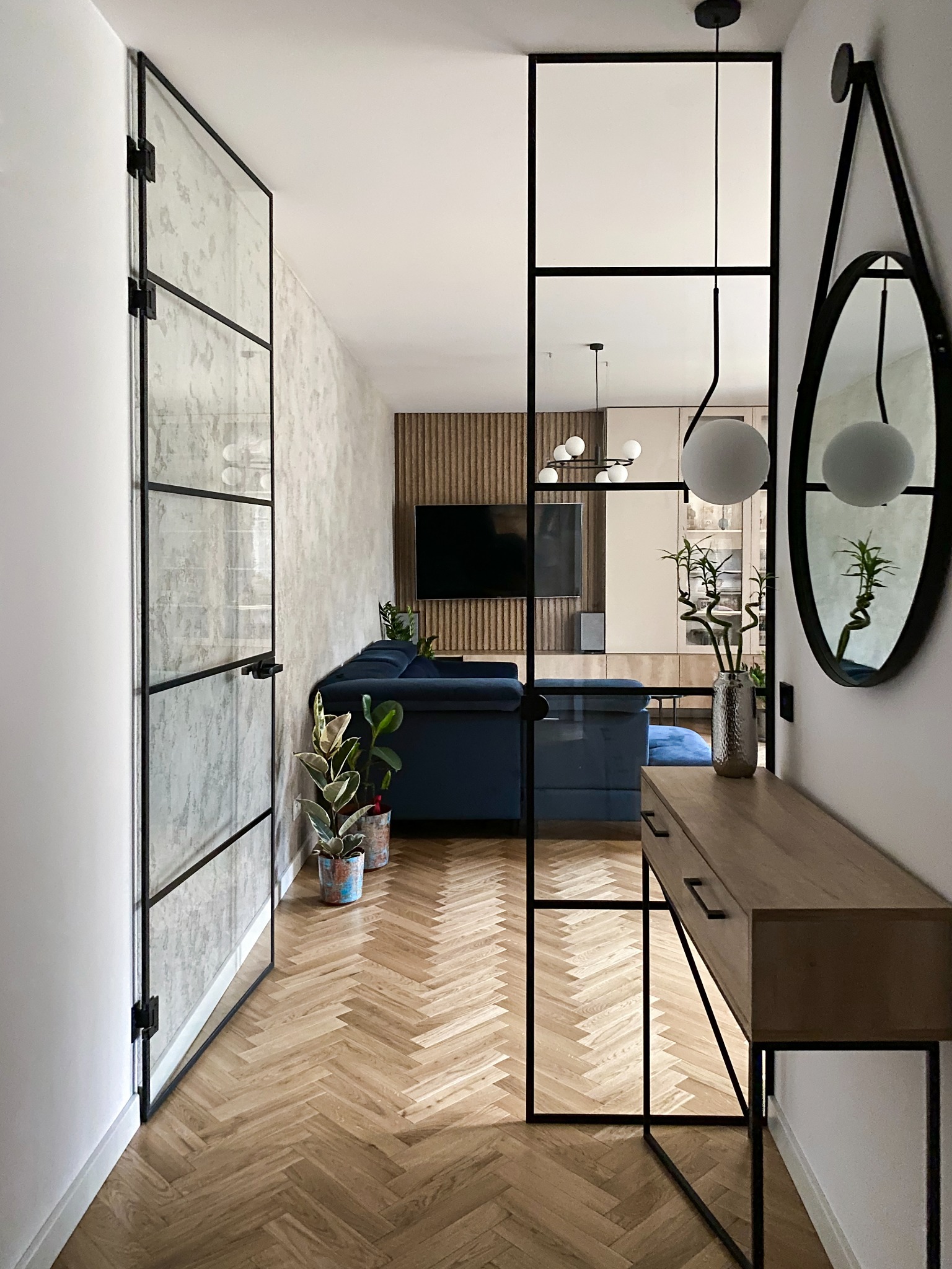 Wnętrze mieszkania z parkietem w jodełkę, szklaną ścianką działową z czarnymi profilami, niebieską sofą i lustrem w czarnej ramie, widok z korytarza.
