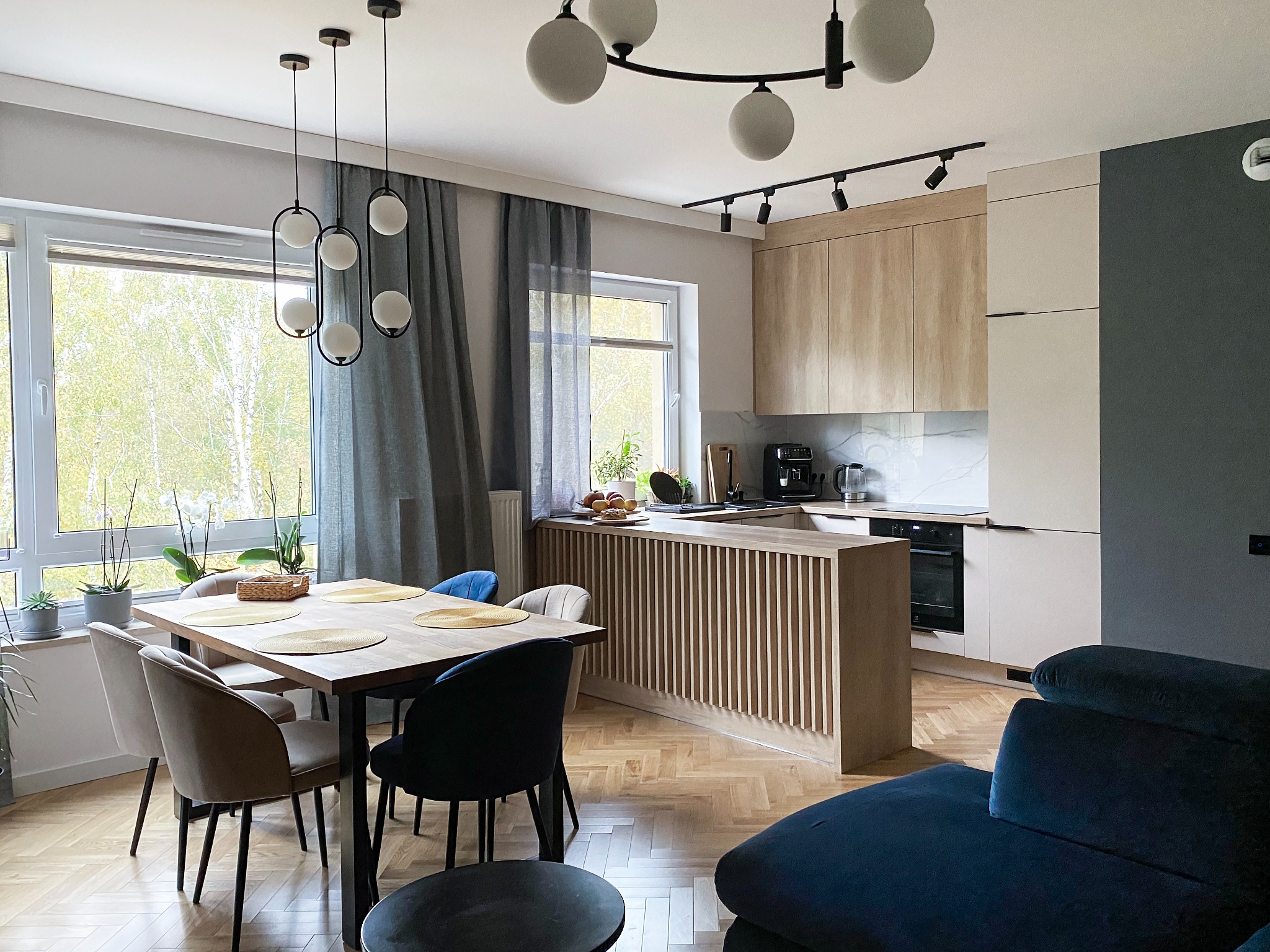 Jasne wnętrze z drewnianą podłogą w jodełkę, stołem z ciemnymi krzesłami i wyspą kuchenną z lamelami, oświetlone nowoczesnymi lampami wiszącymi i reflektorami.