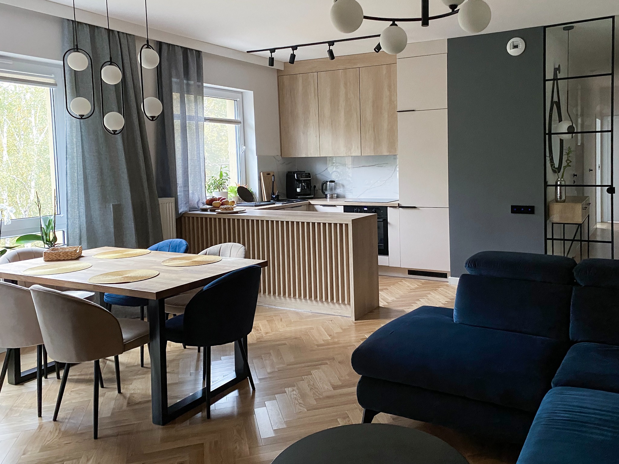 Jasne, przestronne wnętrze mieszkania z parkietem w jodełkę, obejmujące kuchnię z drewnianymi szafkami i białym blatem, stół z krzesłami w odcieniach beżu i granatu oraz ciemnoniebieski narożnik