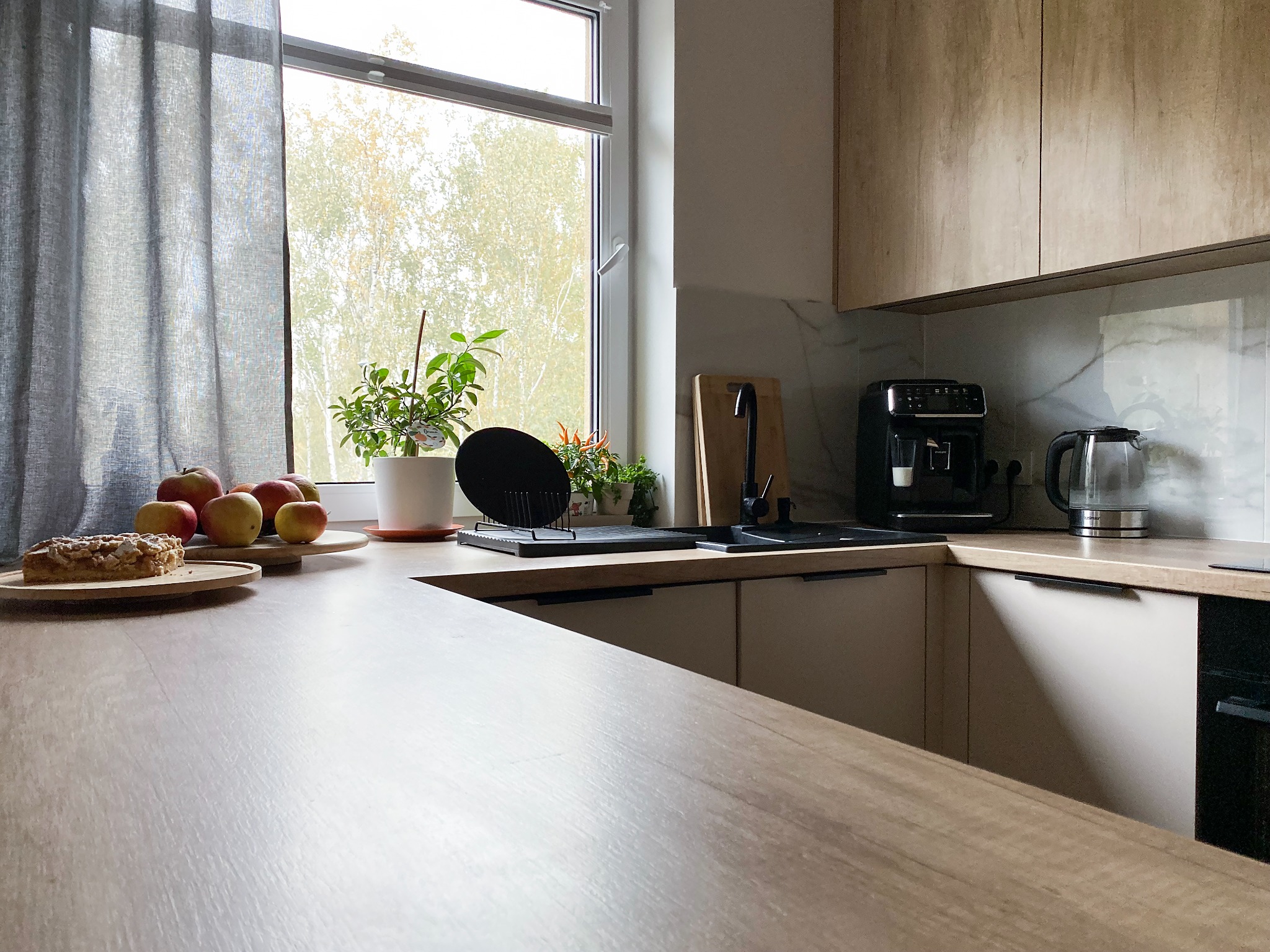 Jasna kuchnia z drewnianymi i beżowymi szafkami, widok na okno z roślinami, jabłka i ciasto na blacie.