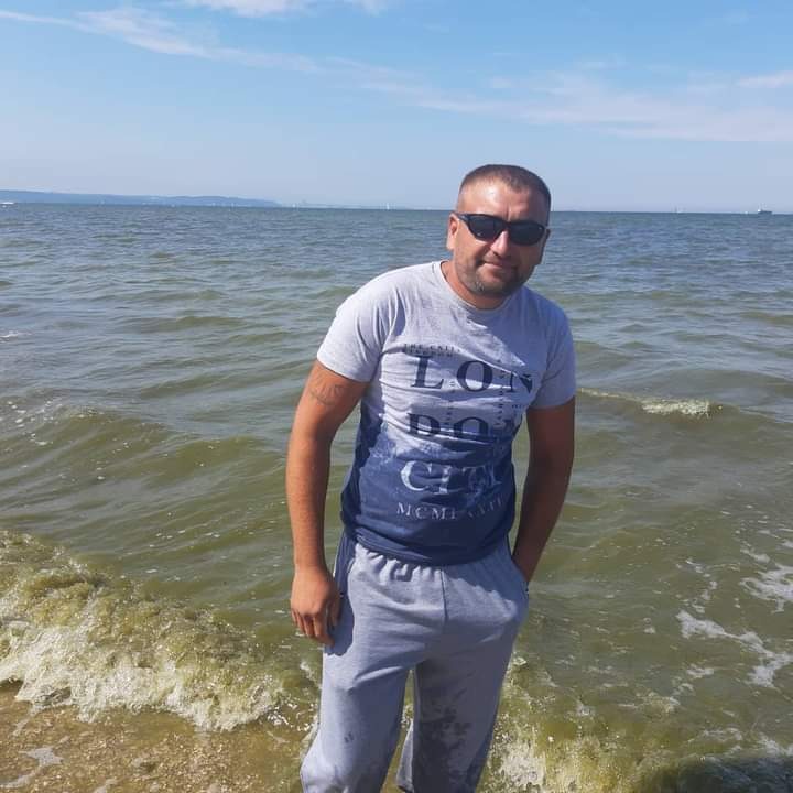 Mężczyzna w okularach przeciwsłonecznych, szarym T-shircie i dresowych spodniach stoi na brzegu morza w słoneczny dzień.