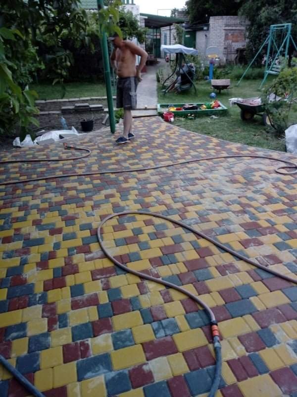 Kolorowa kostka brukowa ułożona na posesji, widoczny wąż ogrodowy i mężczyzna bez koszulki w tle, teren z elementami placu zabaw.