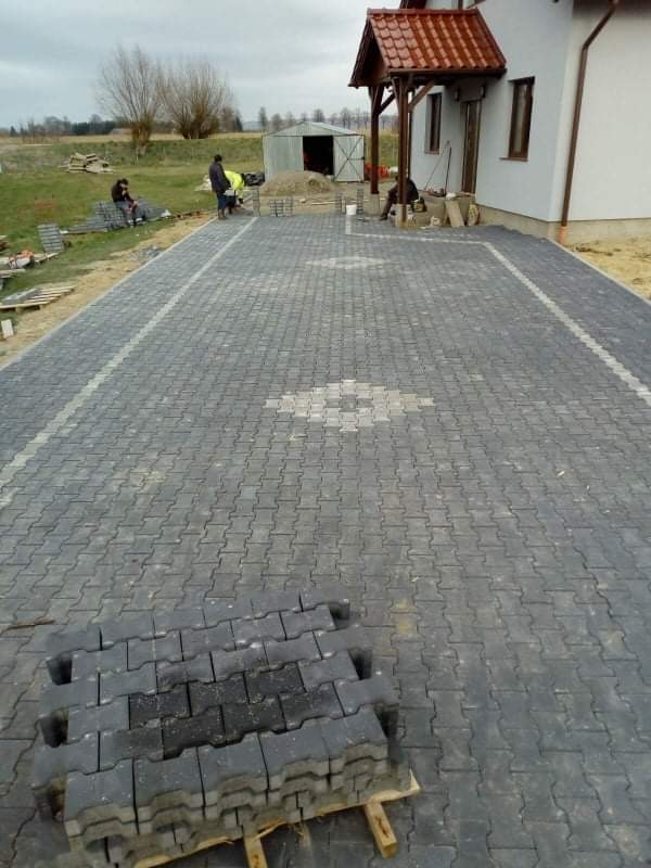 Ułożona nawierzchnia z szarej kostki brukowej przed domem, z widocznym wzorem dekoracyjnym na środku i stosem nieużytej kostki na palecie w pierwszym planie. Prace w toku, widoczni pracownicy...