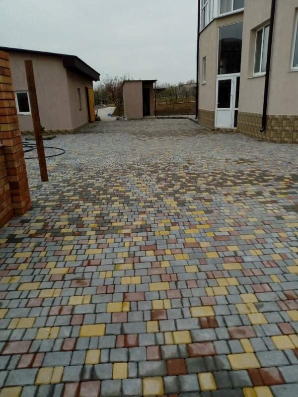 Podjazd z wielobarwnej kostki brukowej ułożonej przed budynkiem mieszkalnym z dobudówką i małym budynkiem gospodarczym, widok w pochmurny dzień.