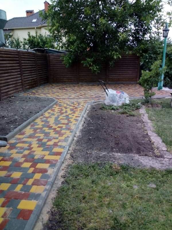 Ścieżka z kolorowej kostki brukowej prowadząca przez ogród z drewnianym ogrodzeniem w tle, widoczny fragment trawnika i przygotowane rabaty kwiatowe.